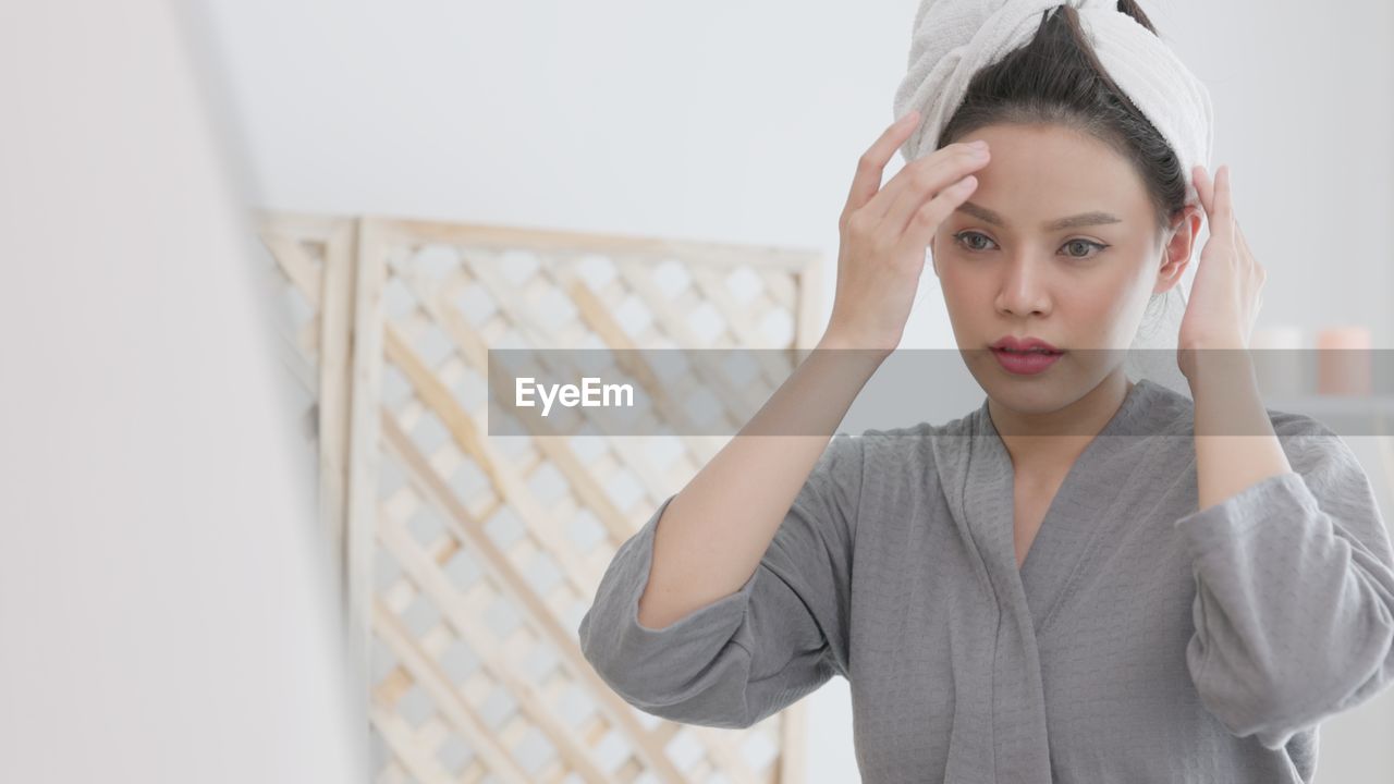 Over a spa background, a young gorgeous woman with a towel on her head cheerfully touches her face.