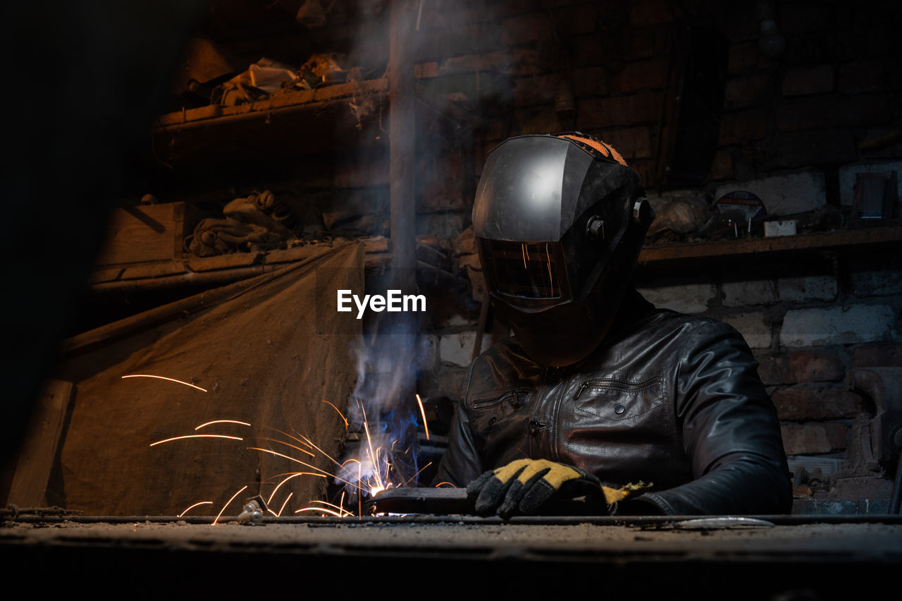 Young mechanic with a welding machine in an old dirty garage at night. man in protection doing