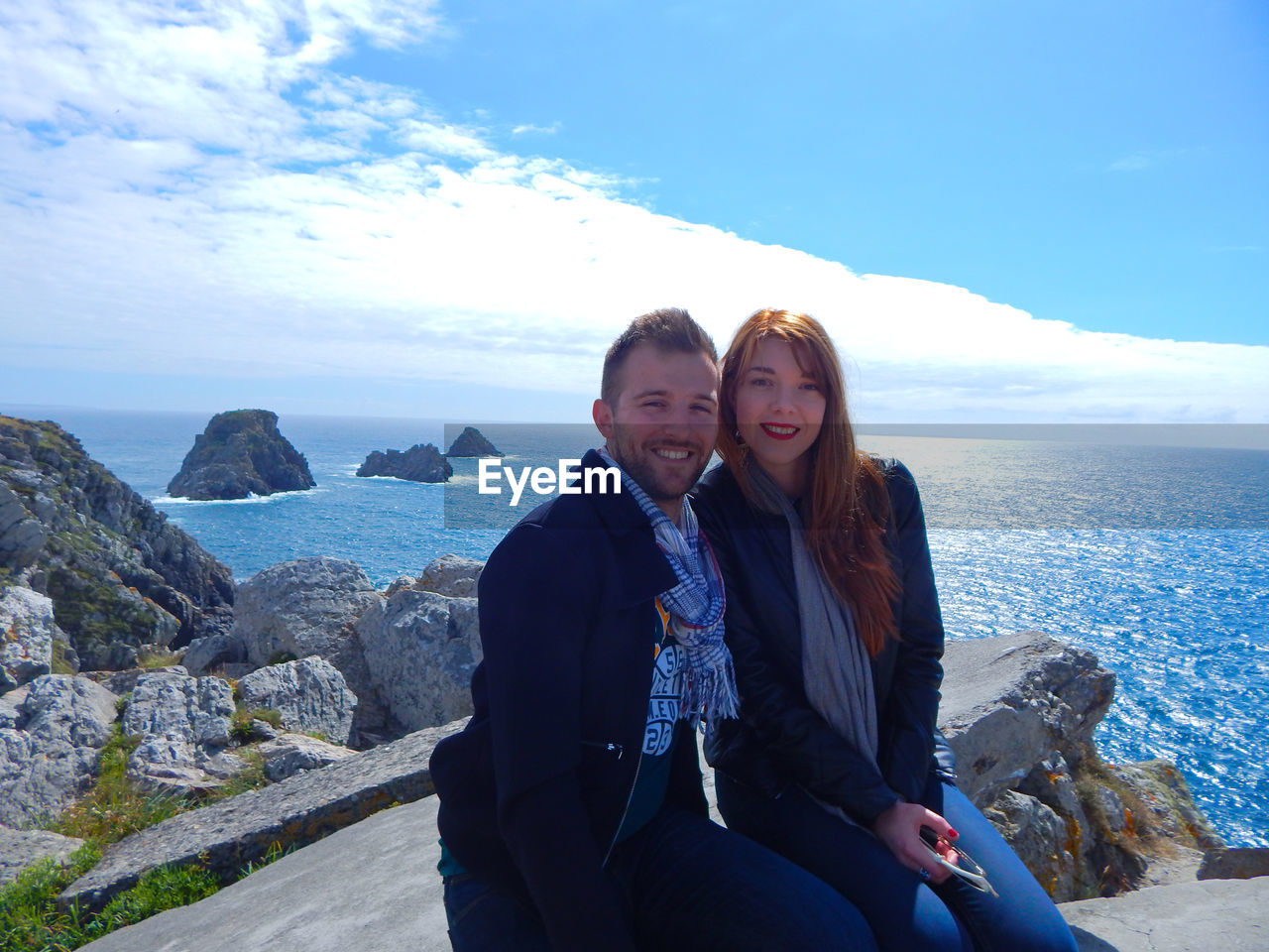 Portrait of smiling couple sitting on rock by sea against sky