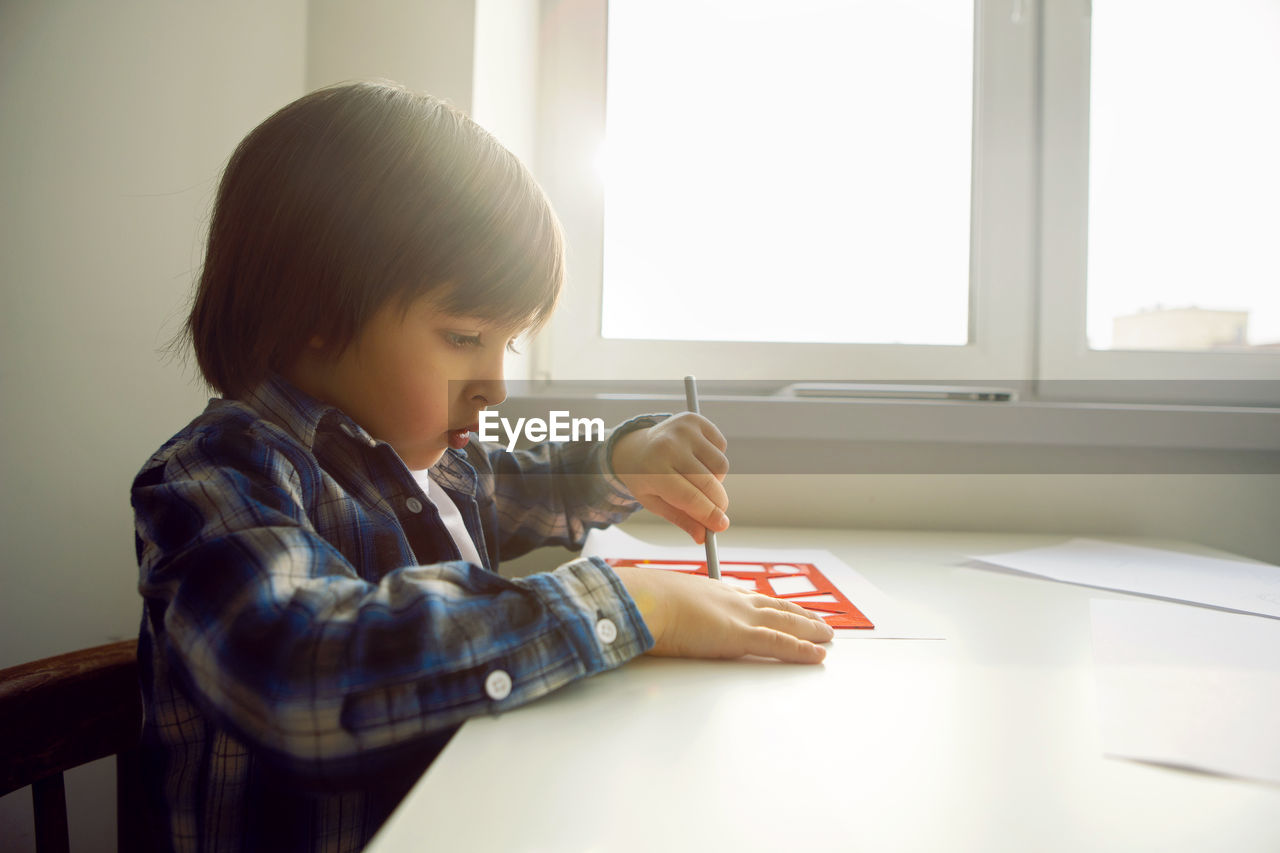 Boy child draws on paper with a ruler on a table sitting by the window