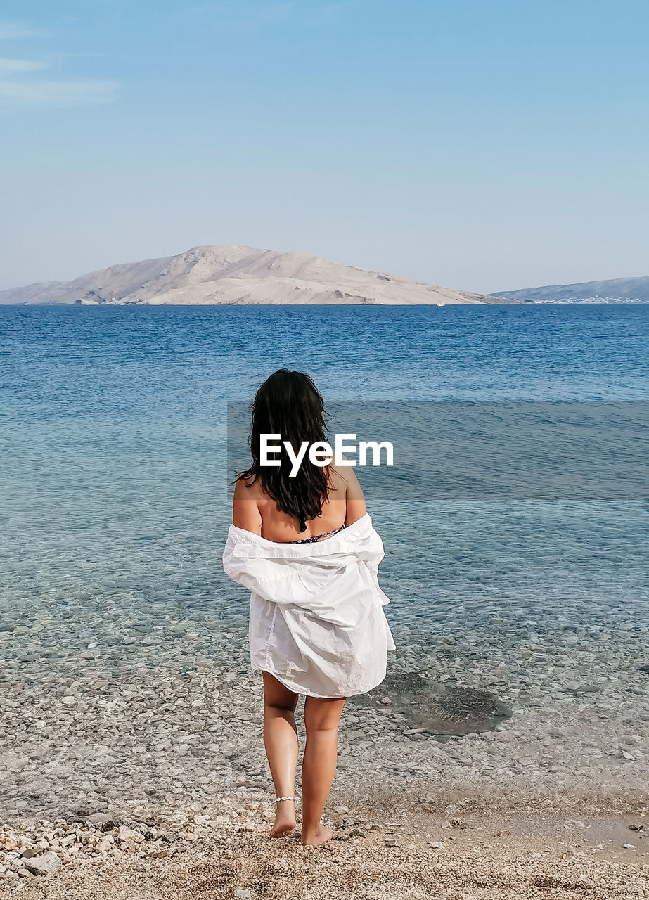 Rear view lifestyle image of stylish young woman in white shirt standing on beach and looking at sea