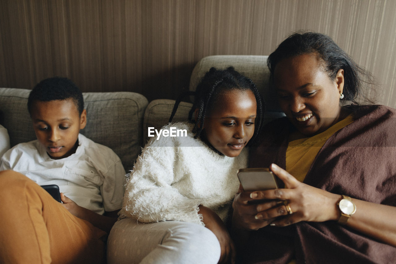 Smiling woman using smart phone while sitting with daughter and son on sofa at home