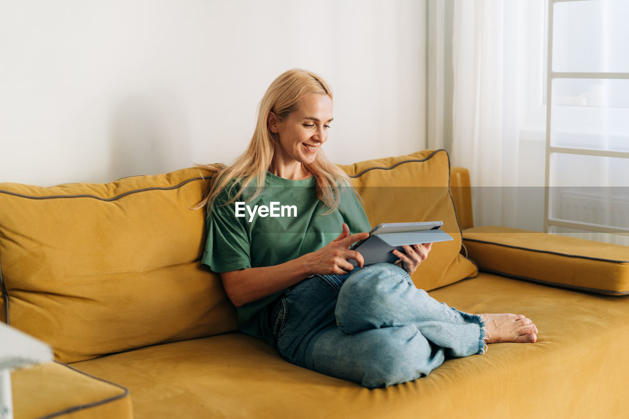 Attractive middle-aged caucasian woman sitting on a sofa works on a computer tablet.