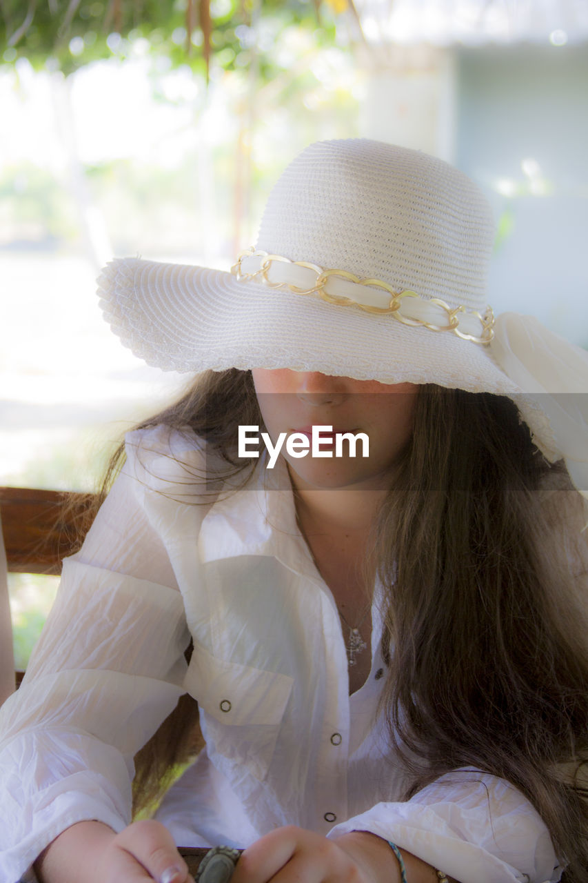 CLOSE-UP OF YOUNG WOMAN WEARING HAT SITTING OUTDOORS