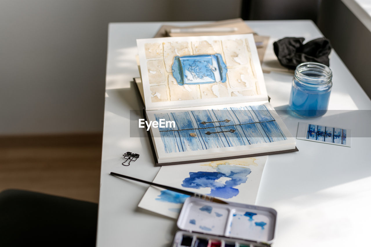 An open watercolor sketchbook lies on a white table in front of the window.