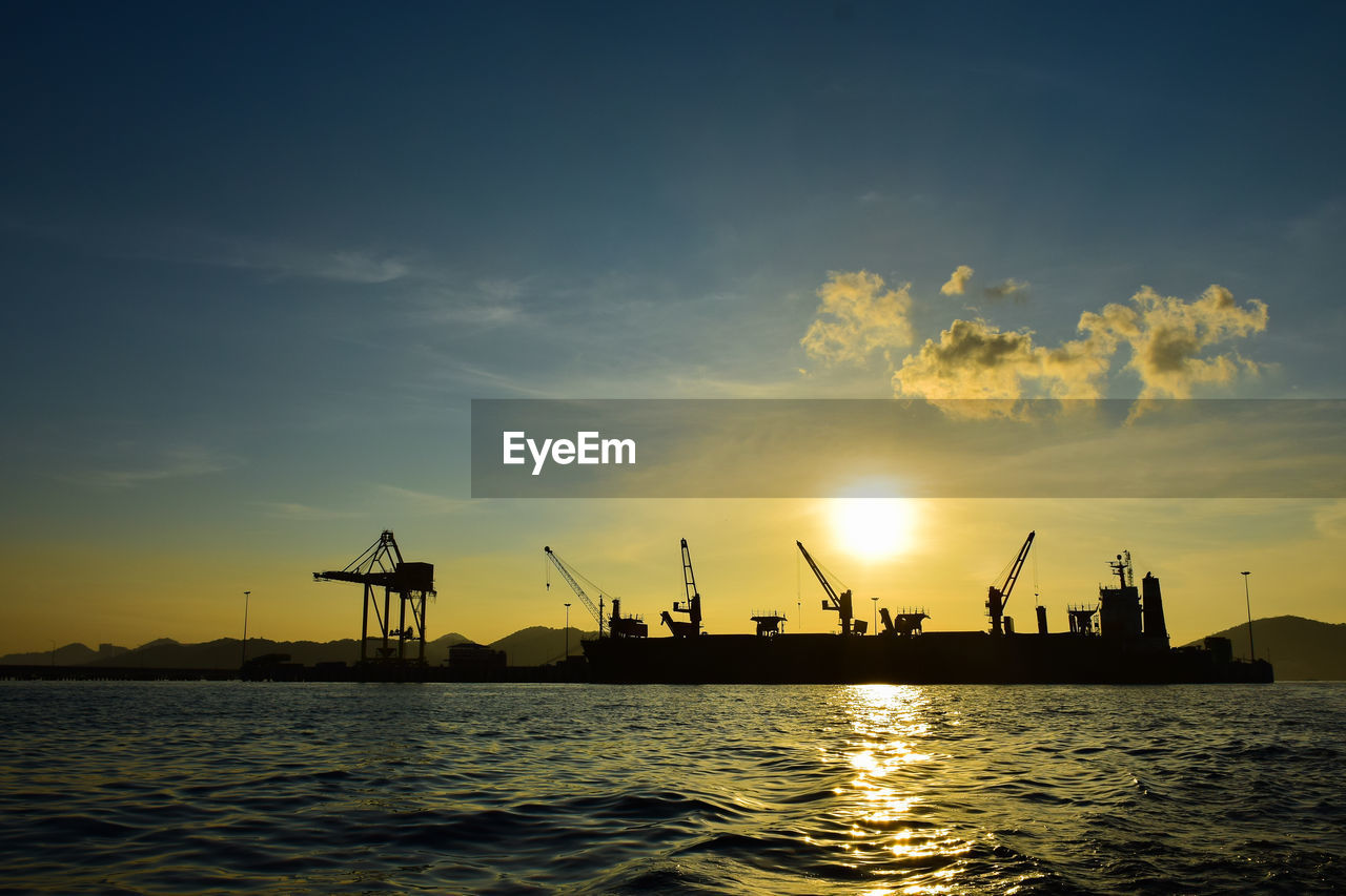 Silhouette cranes at harbor against sky during sunset