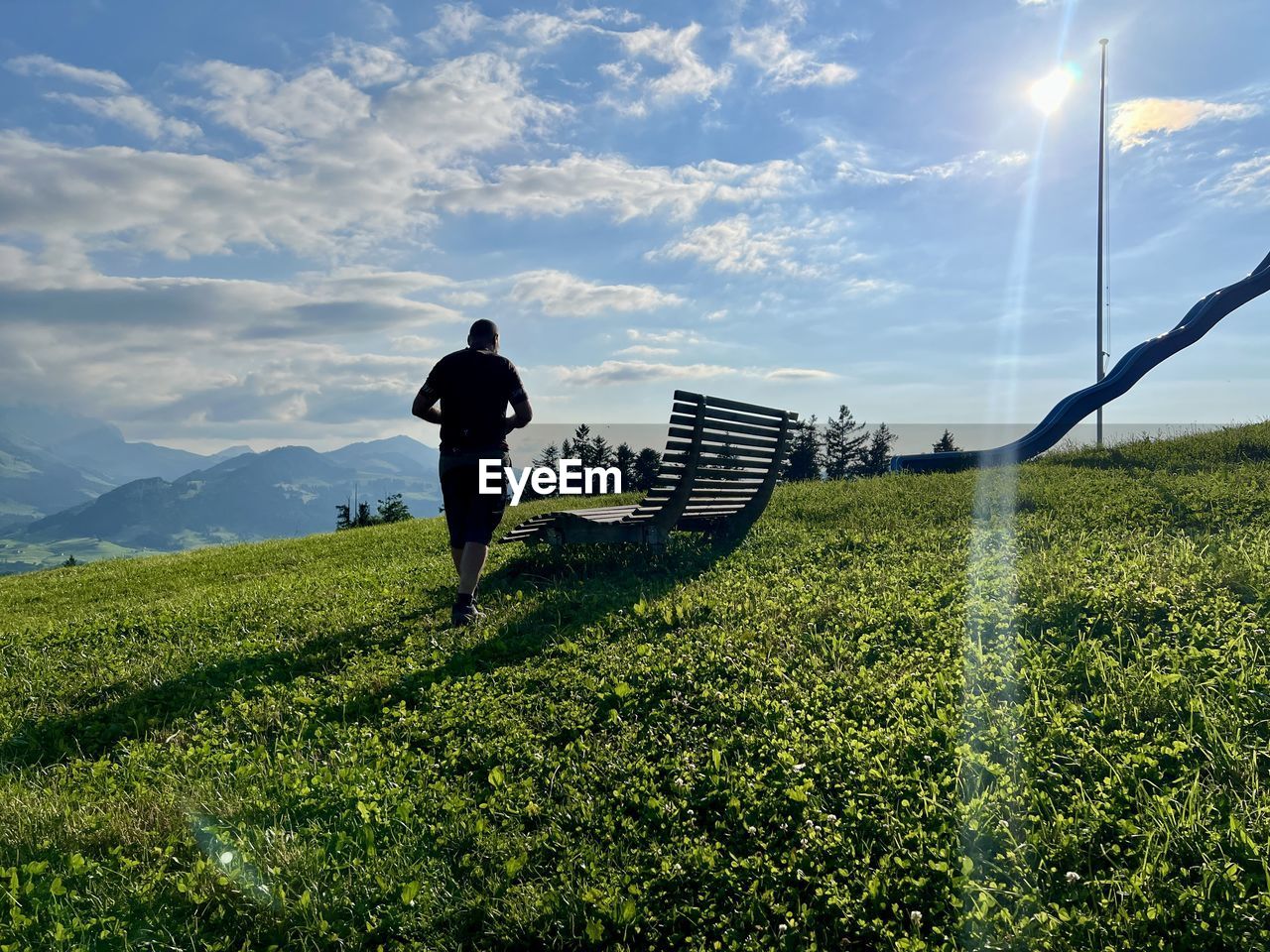 sky, grass, agriculture, field, sunlight, nature, horizon, one person, hill, environment, plant, rural area, landscape, adult, green, cloud, flower, land, meadow, rear view, beauty in nature, men, full length, rural scene, growth, scenics - nature, day, outdoors, leisure activity, lifestyles, occupation, morning, tranquility, grassland, standing, tree, wind, environmental conservation