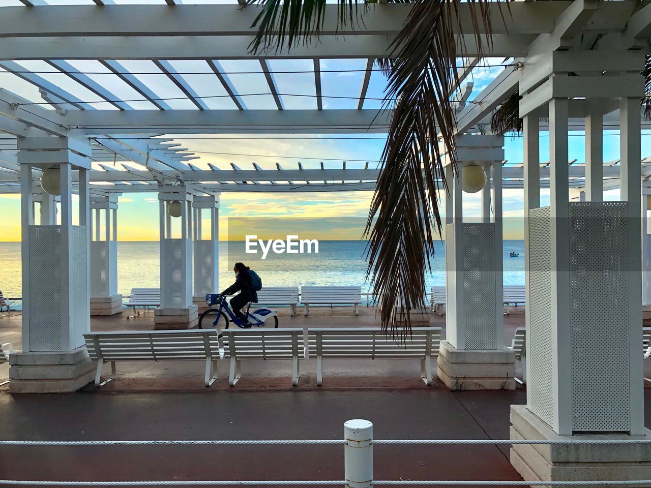 Side view of person riding bicycle on footpath by sea at sunset