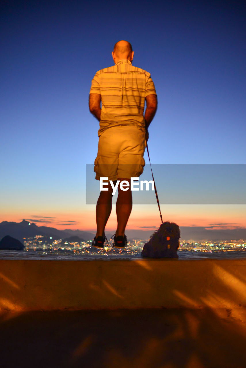 Rear view of man jumping by dog on retaining wall during sunset