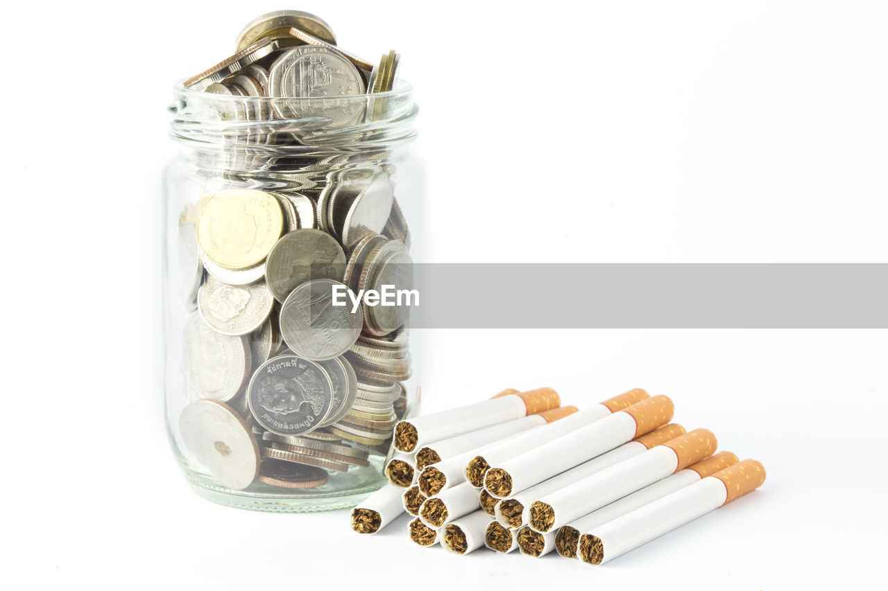 Close-up of coins in jar with cigarettes against white background