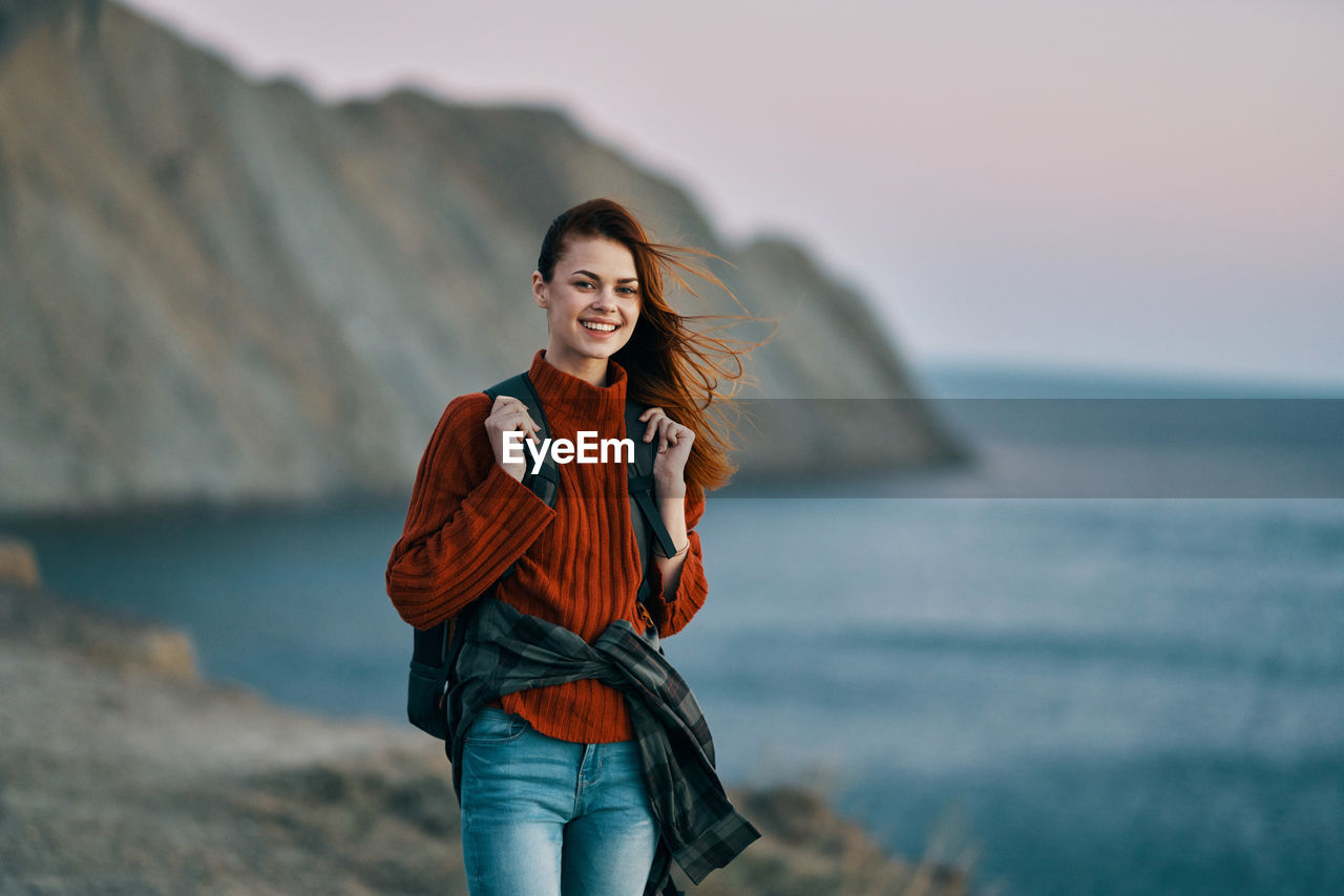 PORTRAIT OF SMILING YOUNG WOMAN STANDING AGAINST SEA