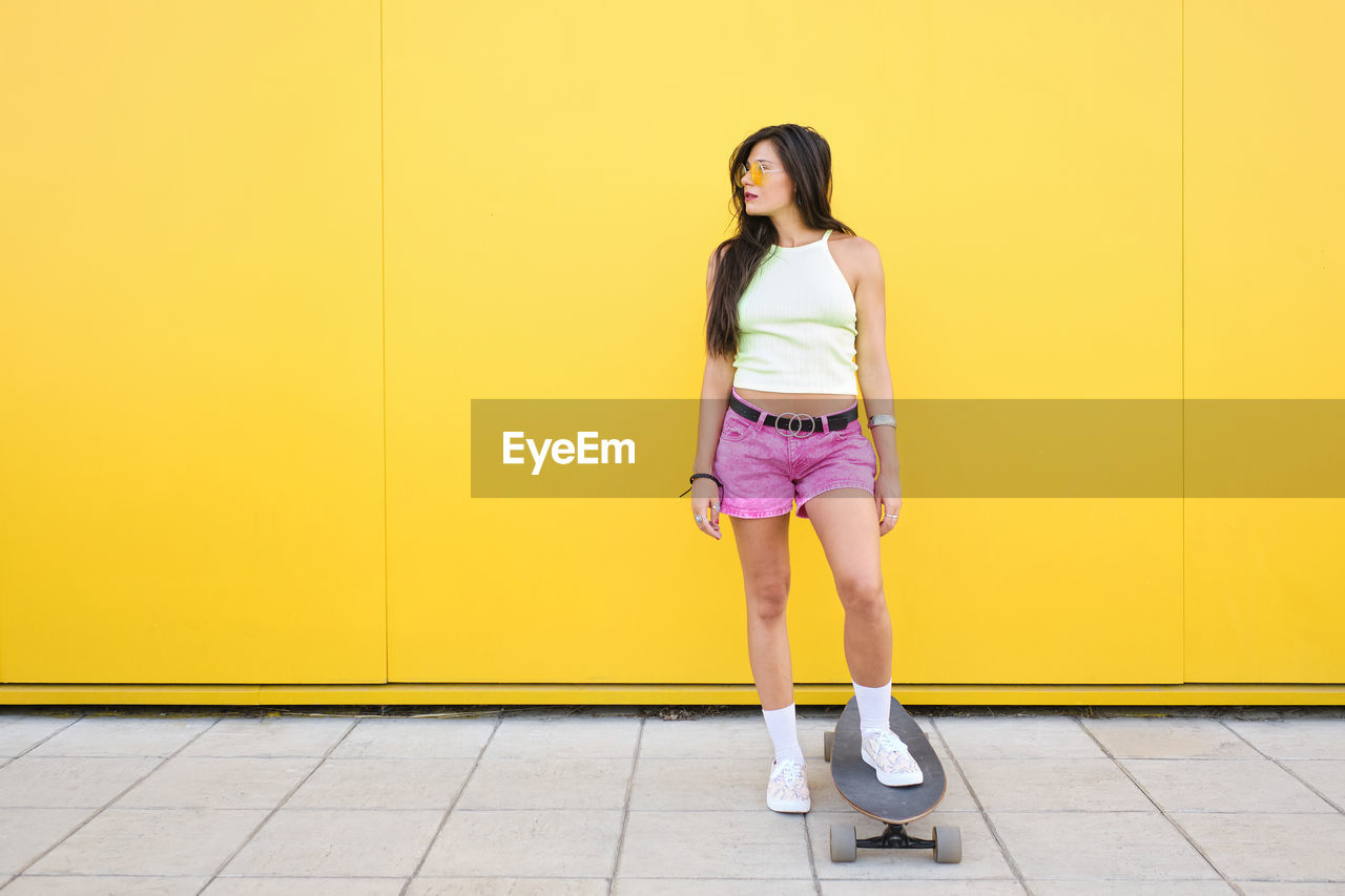 Portrait of beautiful girl standing with skateboarding in front of yellow wall