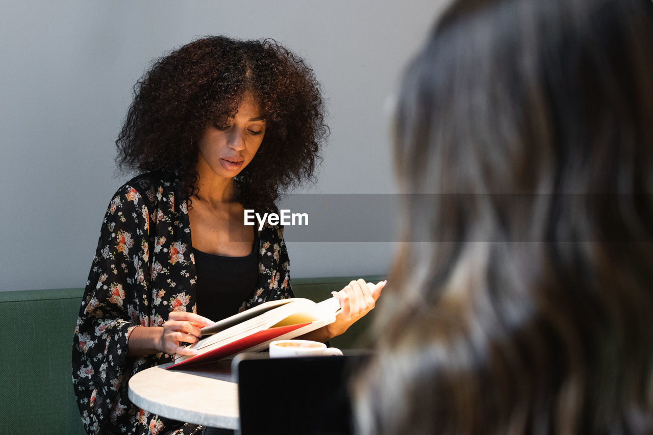 Young ethnic female with afro hairstyle reading textbook at cafeteria table against crop unrecognizable friend