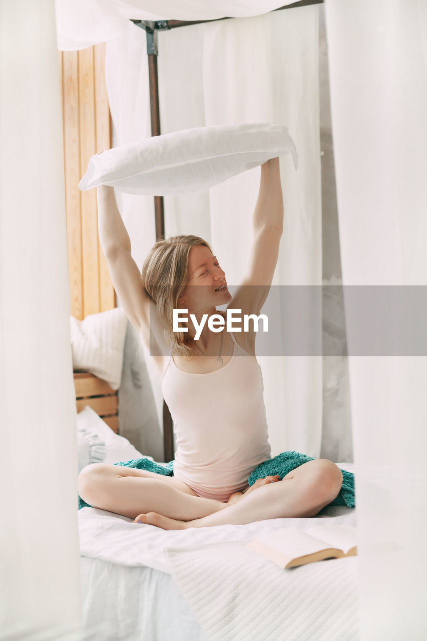 A beautiful smiling girl sits cross-legged on the bed and holds a pillow on her outstretched arms. 