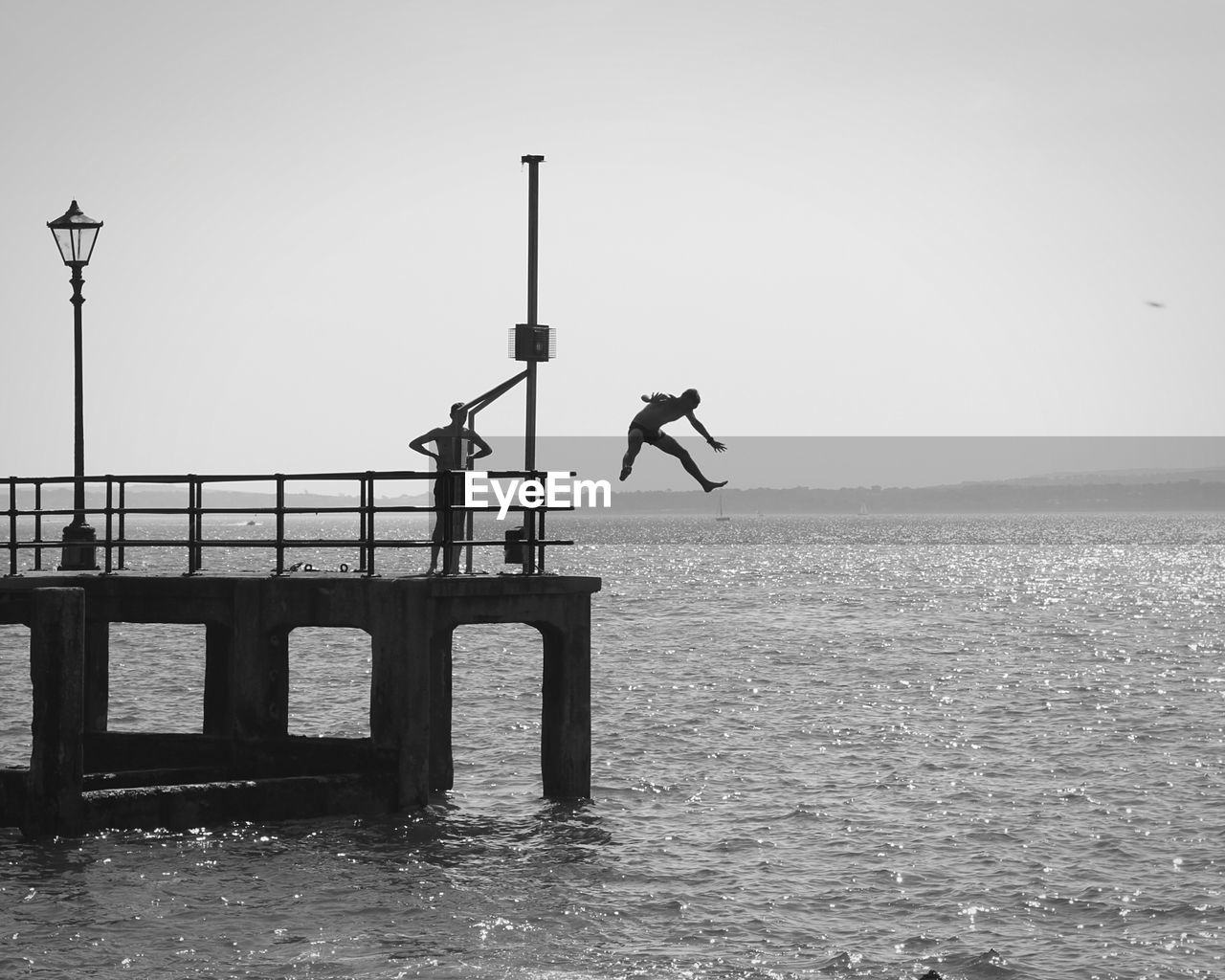 Man standing on pier while looking at friend jumping in sea against clear sky