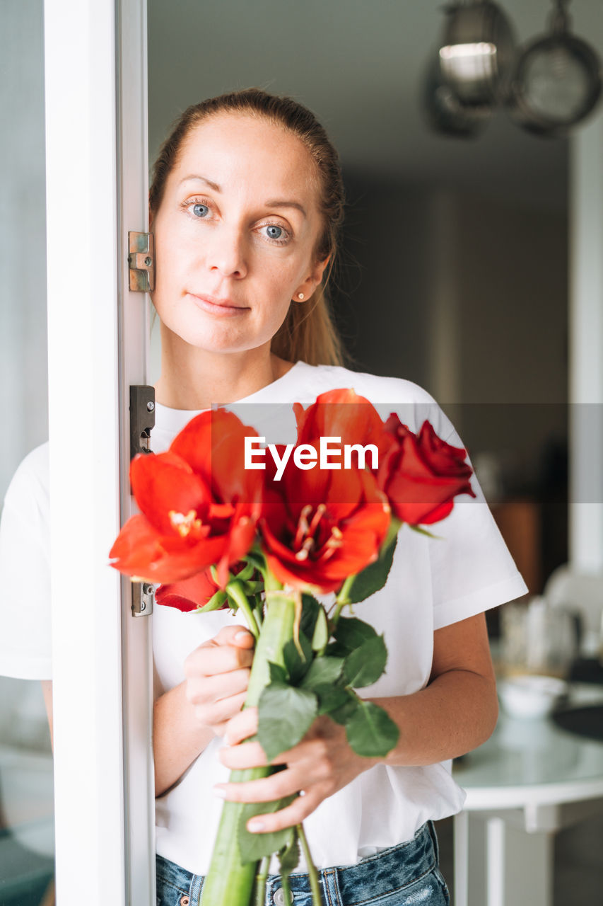 Portrait of young woman in white t-shirt with bouquet of red flowers in hands near window in kitchen 