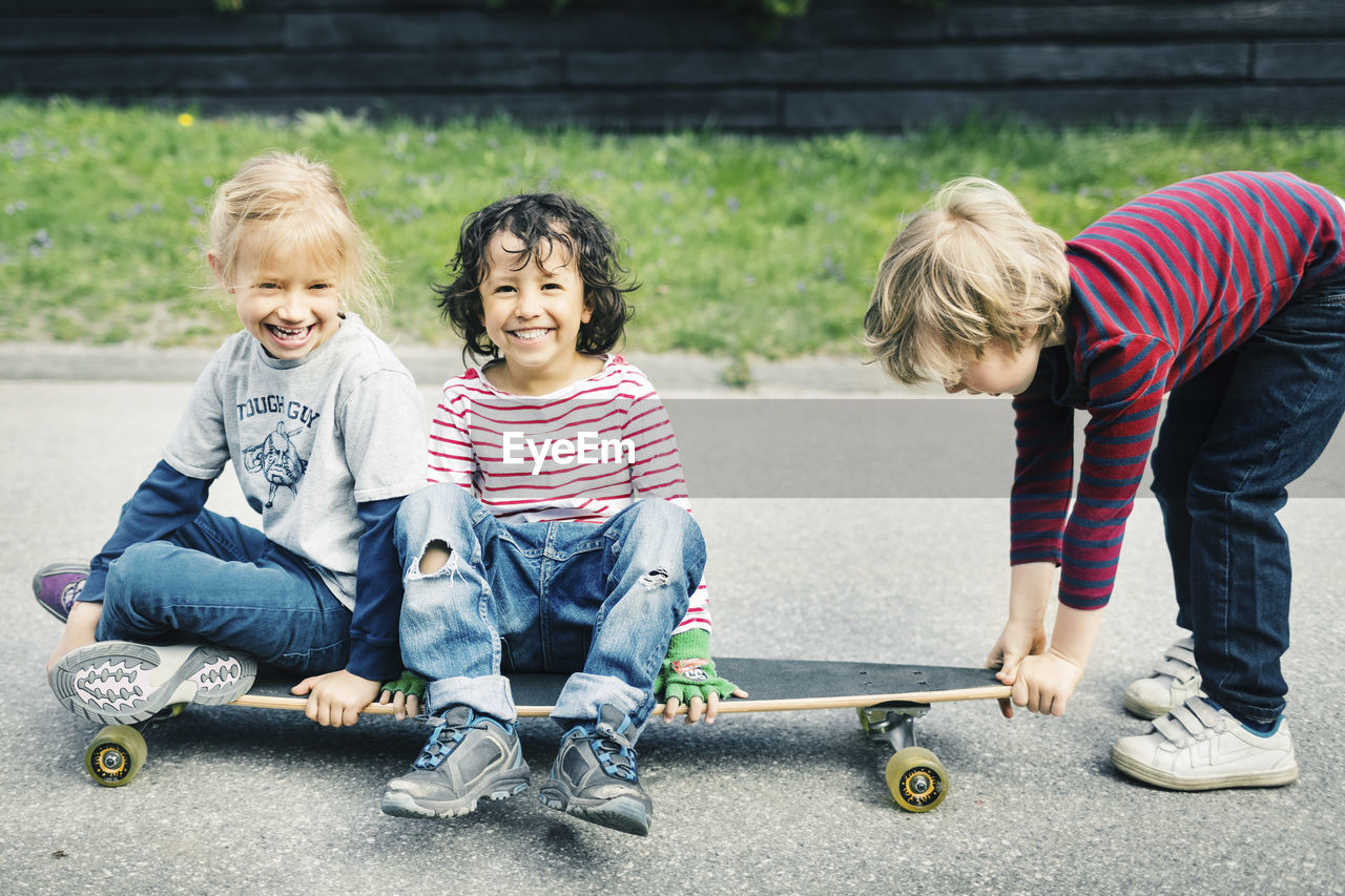 Portrait of happy children sitting on skateboard while friend pulling it at yard