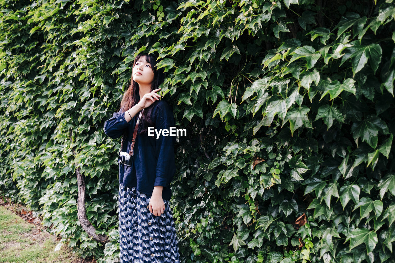 Young female photographer with retro style camera looking up standing by plants