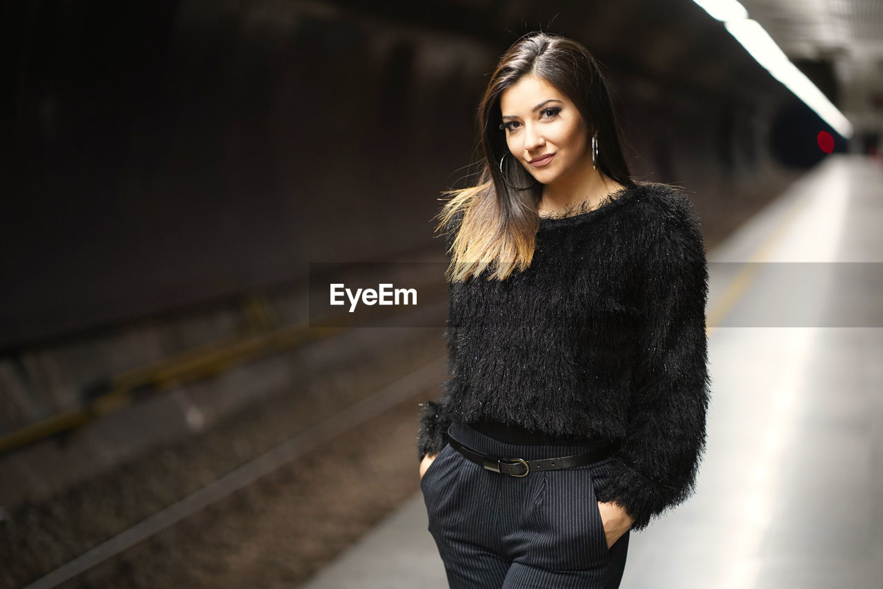 PORTRAIT OF SMILING YOUNG WOMAN STANDING AGAINST BLURRED BACKGROUND