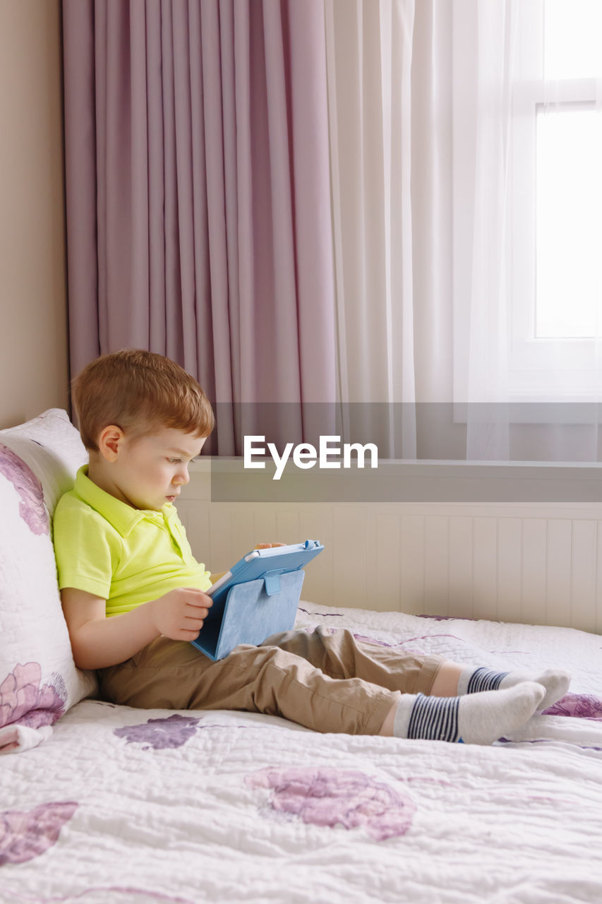 Cute boy using digital tablet while sitting on bed at home