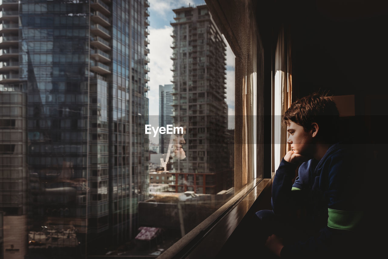 Tween boy looking out a window at tall buildings of the city outside.