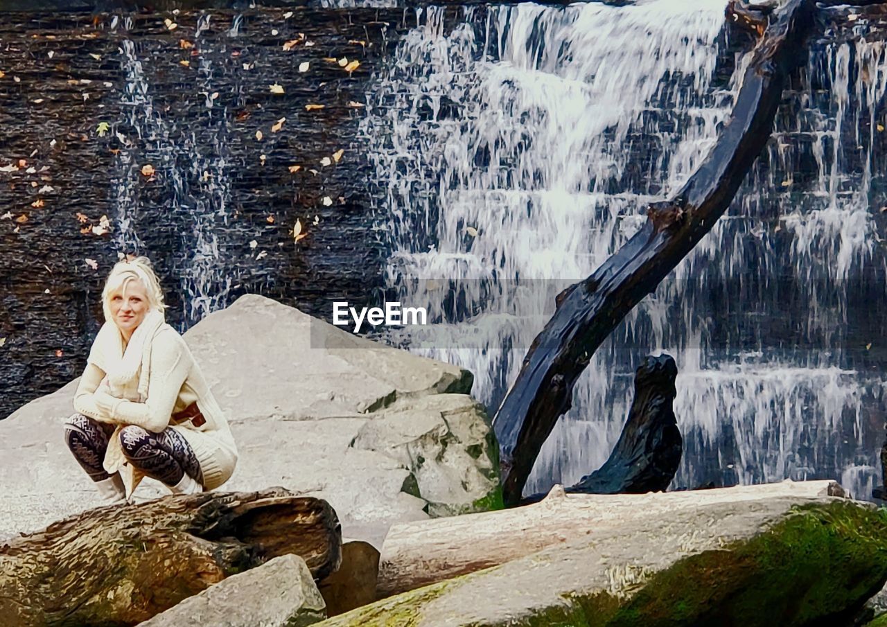 PORTRAIT OF WOMAN SITTING ON ROCK AT WATERFALL