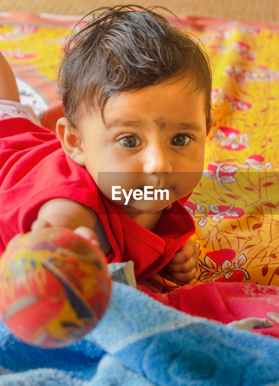 A cute indian baby holding a ball towards the camera while lying down on a bed-sheet over a mat.