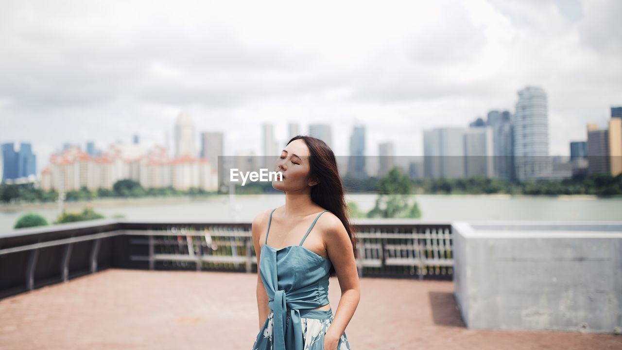 Young woman with eyes closed standing against cityscape and sky