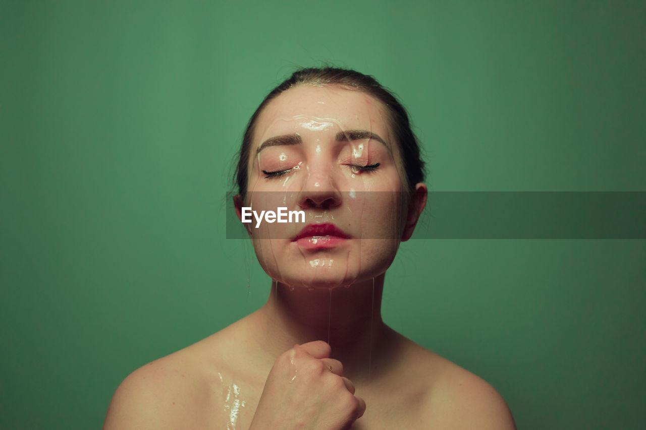 Close-up of shirtless young woman with beauty product on face against green background