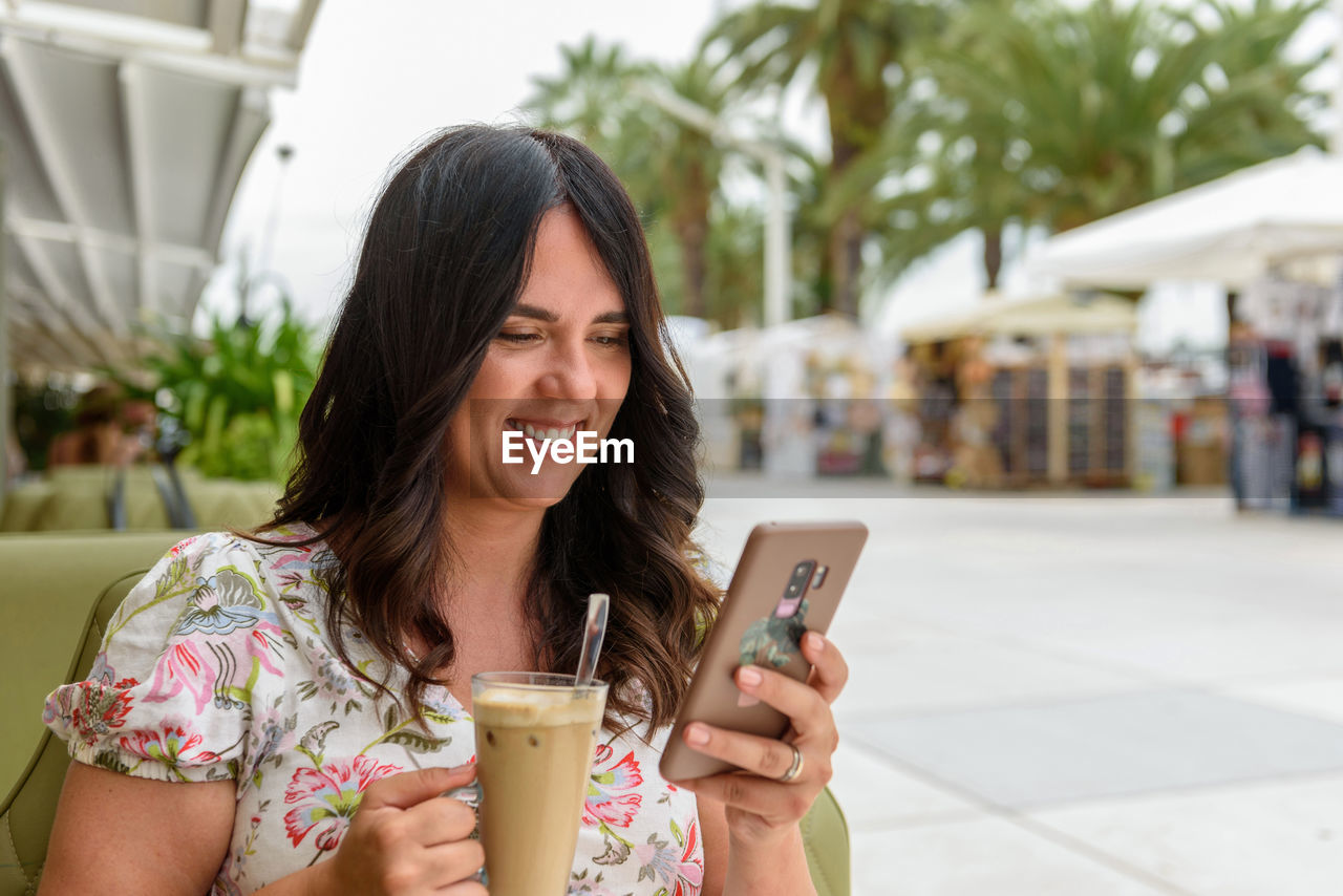 Pretty girl with long hair sitting in bar outdoor in city, using phone and drinking iced coffee