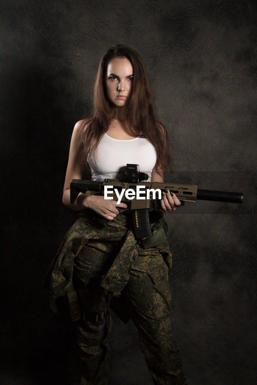 Portrait of beautiful young woman holding gun while standing against wall