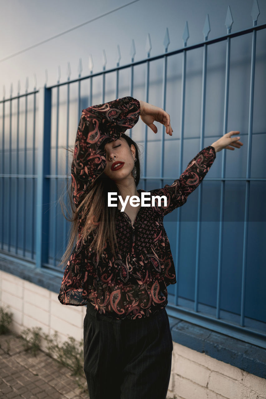 Stylish young ethnic female with long dark hair in trendy dress standing against blue wall on street and closed eyes thoughtfully