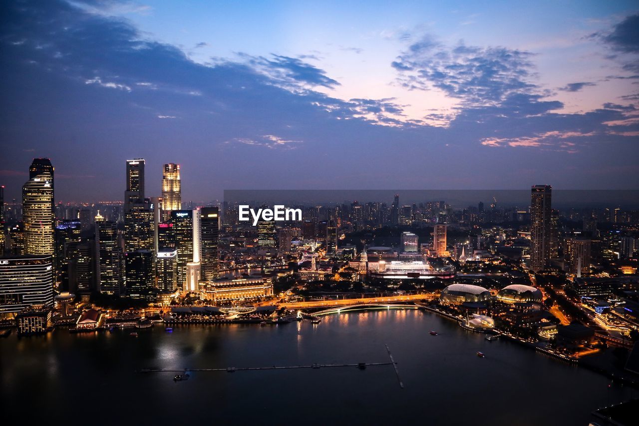 Illuminated buildings by river against sky in city