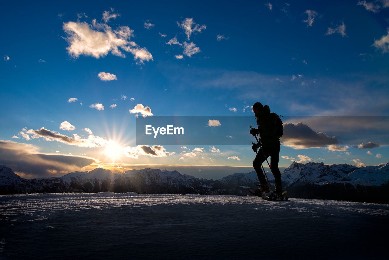 MAN STANDING ON SNOW COVERED MOUNTAIN AGAINST SKY DURING SUNSET