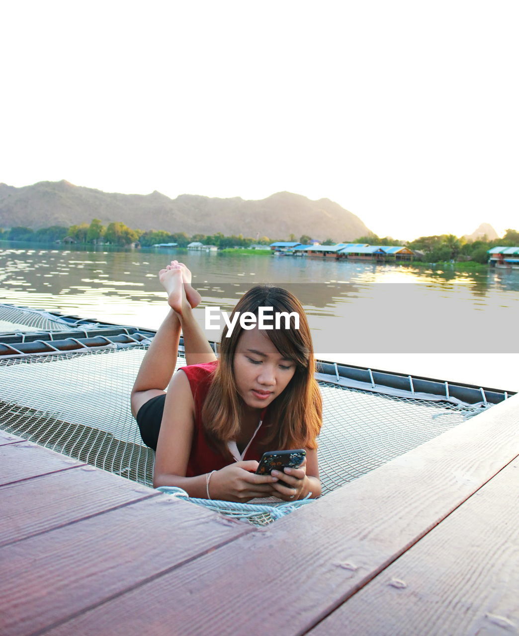 Woman using phone while lying on net by lake