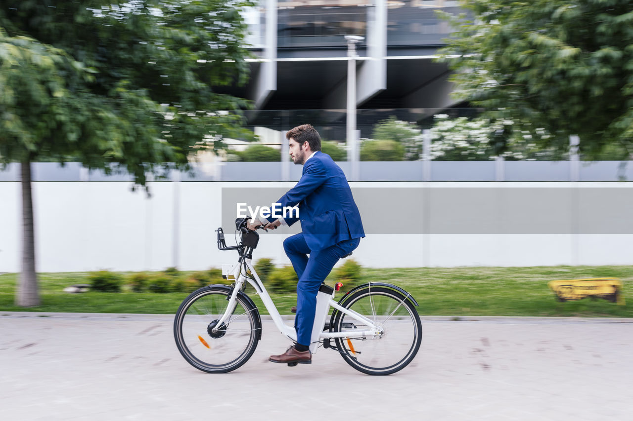 Young smiling man in elegant suit riding electric bicycle on pavement while looking away