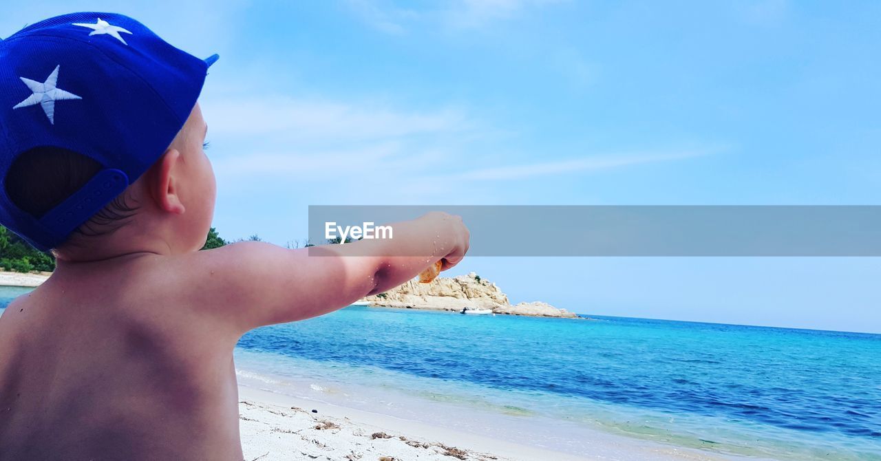 Shirtless baby boy pointing at sea against sky