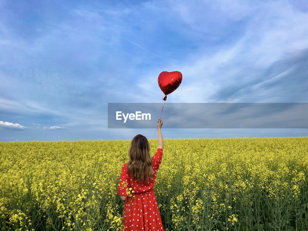 Woman in red dress standing in a field of canola flowers and holding a red heart balloon