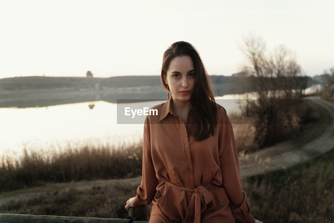 Portrait of young woman standing by lake against sky