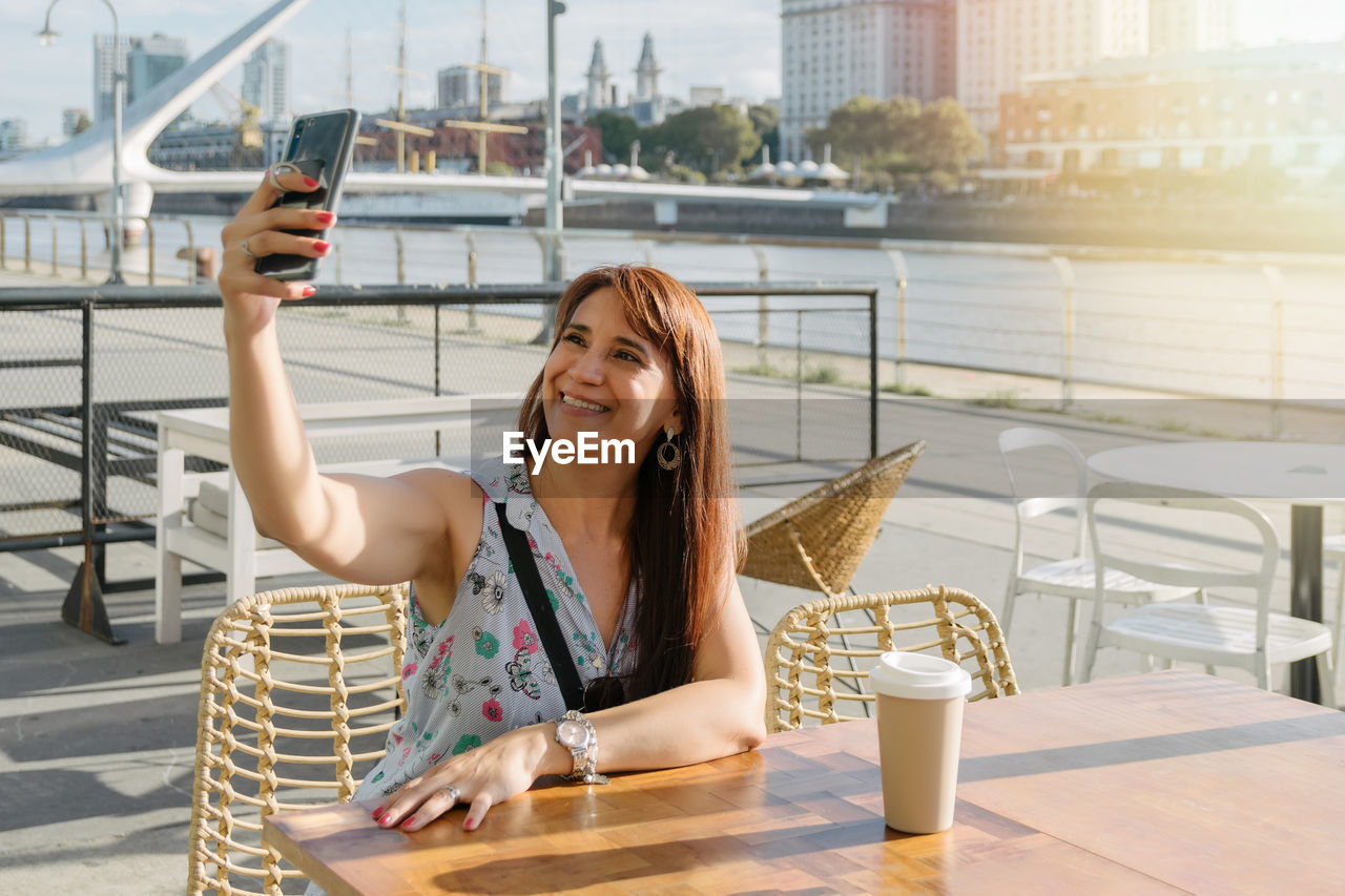A hispanic woman taking a selfie with her phone on coffee shop  outside drinking coffee.