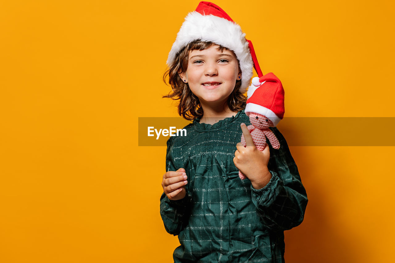 Cute girl in green dress and santa hat standing looking at camera with crochet toy during christmas celebration against yellow background