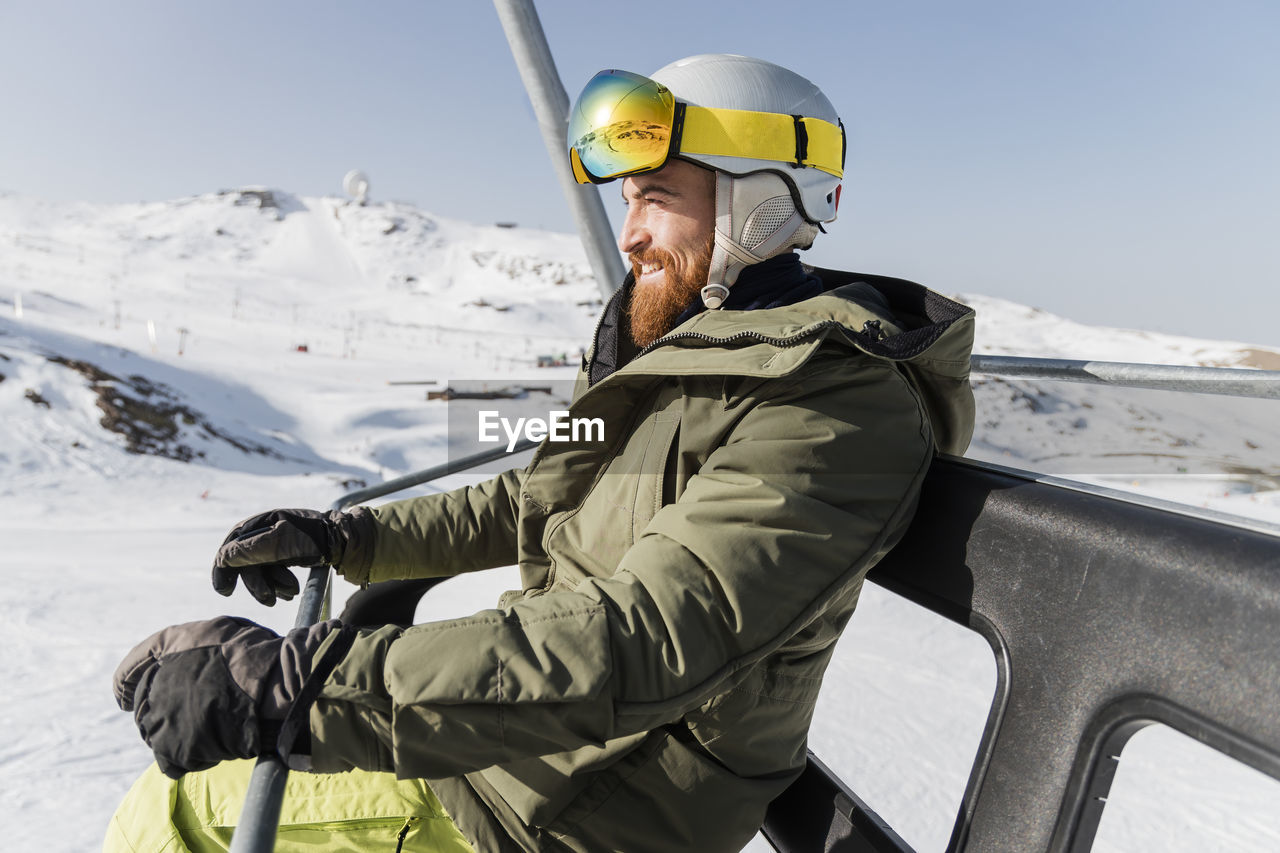 Young man wearing ski helmet and goggles sitting on lift