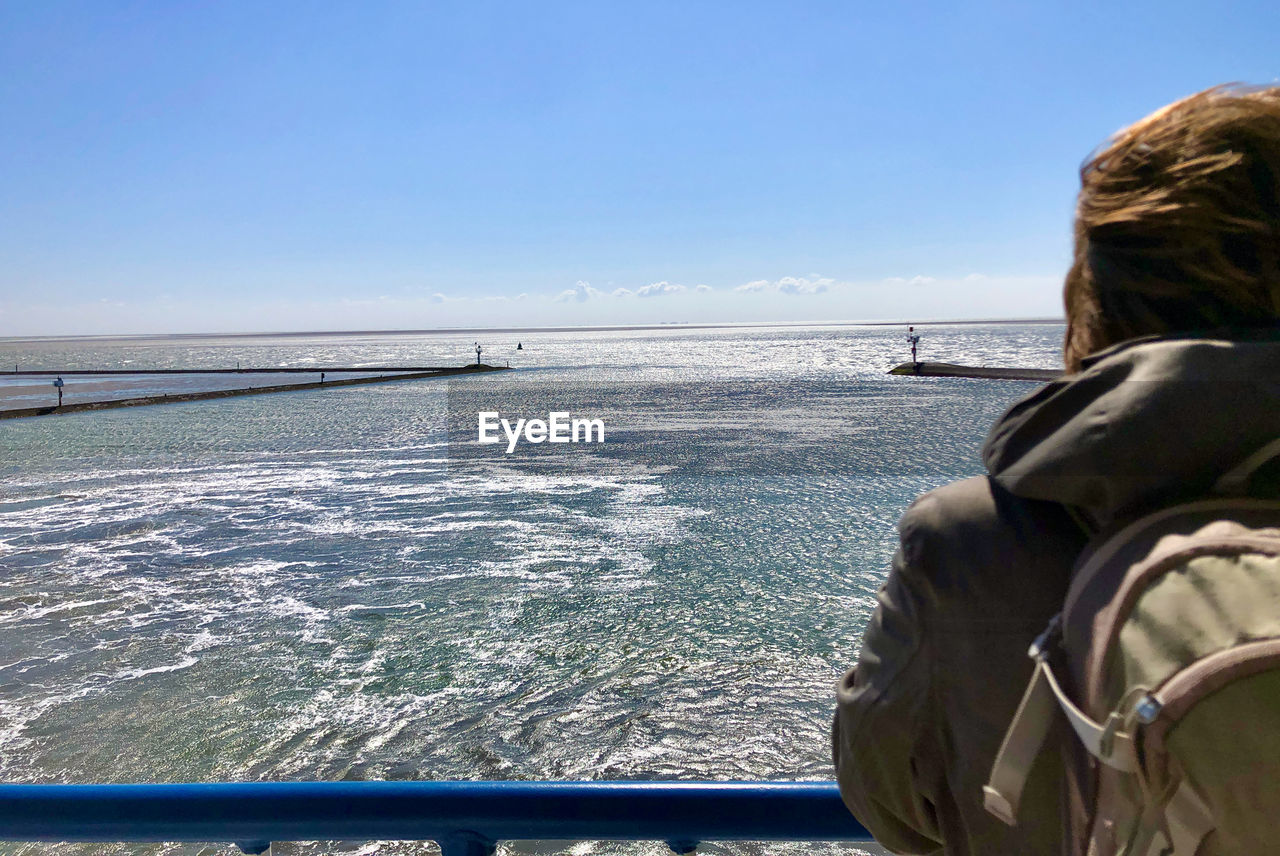 Partial rear view of woman leaning on the railing of a ship, watching the sea under blue sky