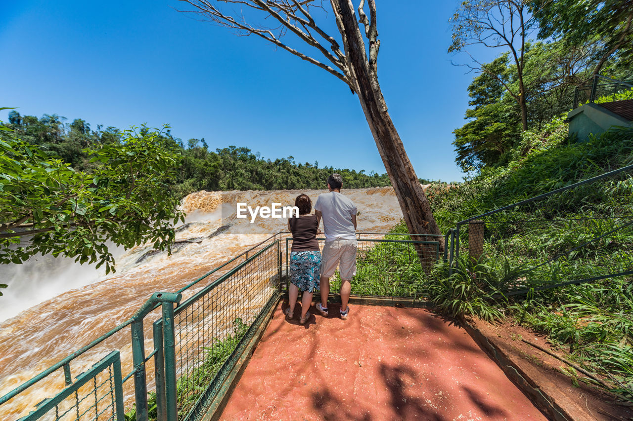 Rear view of couple standing by railing against waterfall