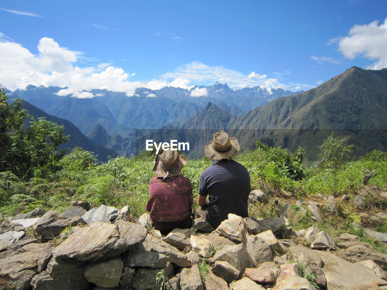 REAR VIEW OF COUPLE SITTING ON ROCK AGAINST SKY