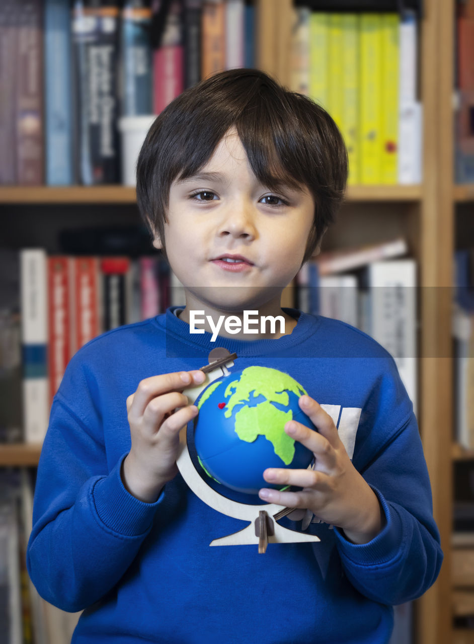 Portrait of boy holding globe against bookshelf