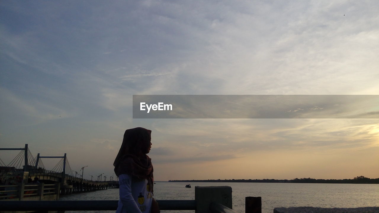 Side view of woman wearing hijab while looking at lake against sky during sunset