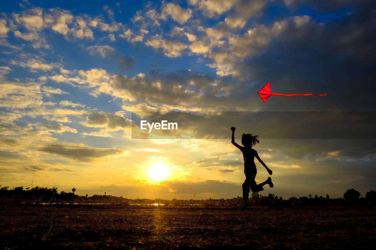 Silhouette girl flying kite while running on landscape during sunset