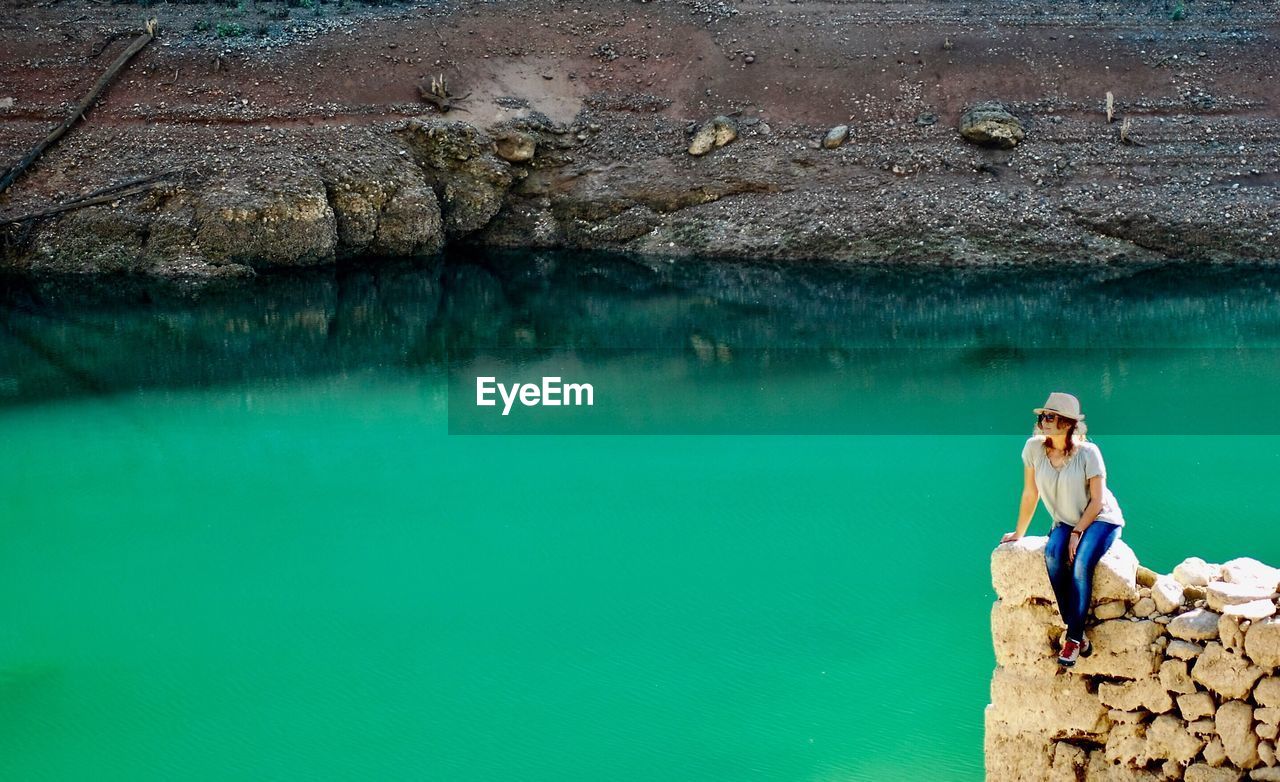 Full length of woman sitting on rock formation by lake against field