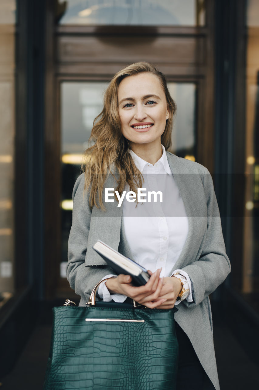 Portrait of smiling businesswoman with book and bag standing by store in city